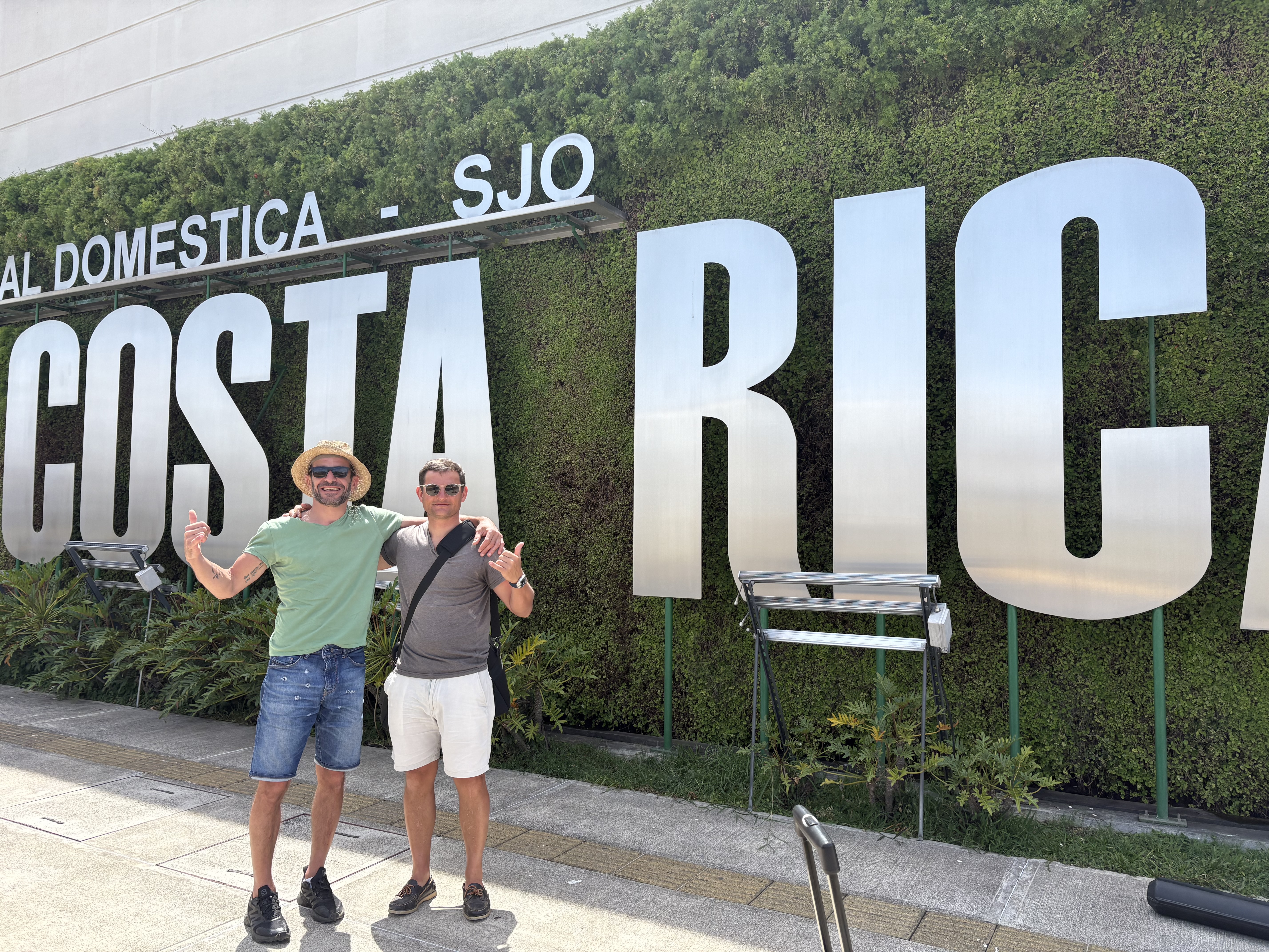 Deux hommes sourient devant un grand panneau COSTA RICA recouvert de verdure, dans un lieu extérieur près de l'aéroport SJO.