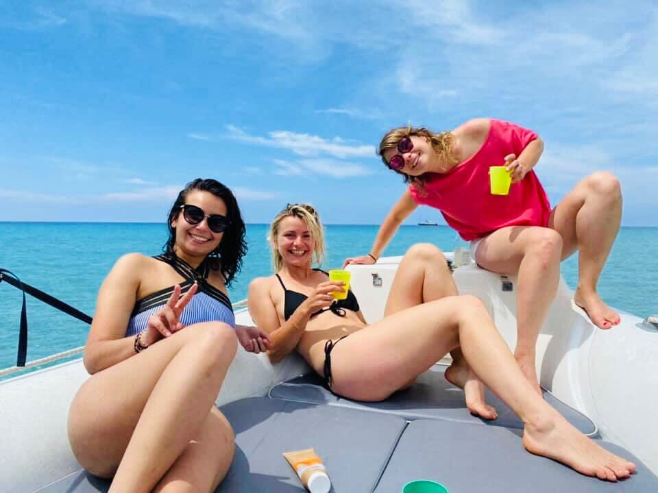 Trois femmes en maillots de bain sur un bateau, tenant des boissons et profitant d'une journée ensoleillée.